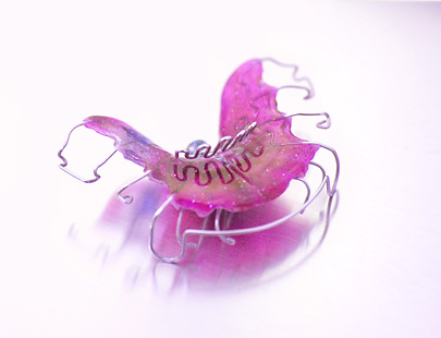 Rosa herausnehmbarer Retainer (zahnärztliche Spange) liegt auf glänzender, weiß-pinker Oberfläche; gebogene Metallbögen umschließen ihn, weiche Schatten und Spiegelung erzeugen eine diffuse Studioatmosphäre.