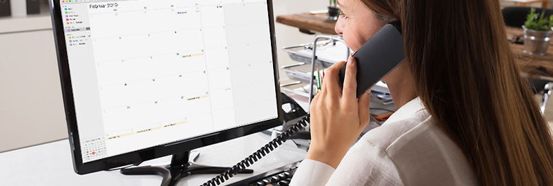 Eine Frau sitzt am Schreibtisch, spricht ins Telefon und blickt auf einen Monitor mit Monatskalender.