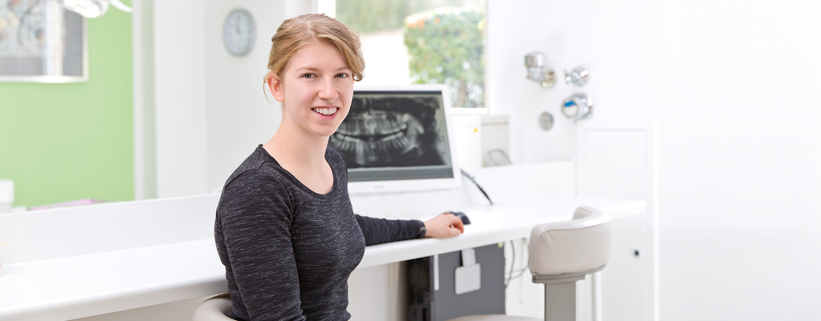 Junge Frau sitzt lächelnd am Schreibtisch, benutzt eine Computermaus vor einem Zahn‑Röntgenbildschirm in einer hellen, modernen Kieferorthopädie.