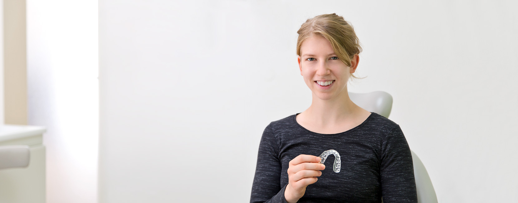 Durchsichtige Zahnschiene (Aligner), die eine lächelnde Frau hält; sie präsentiert sie sitzend in einem hellen, weißen Behandlungszimmer einer Kieferorthopädie.