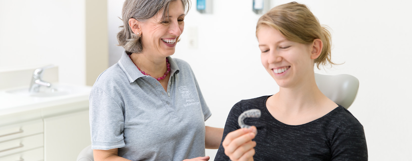Junge Frau hält eine transparente Zahnschiene, lächelt und betrachtet sie, während eine lächelnde Kieferorthopädin neben ihr in einer hellen, modernen Praxis steht.