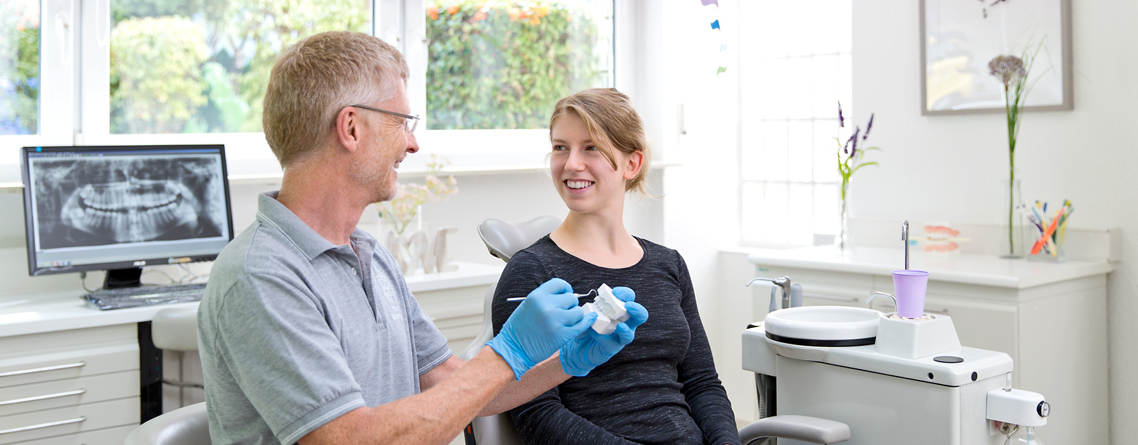 Dr. Süßenberger hält Zahnmodell und erklärt es, jugendliche Patientin lächelt im Behandlungsstuhl; helle Kieferorthopädie mit Zahn-Röntgenbild, Instrumenten und Fenster im Hintergrund.