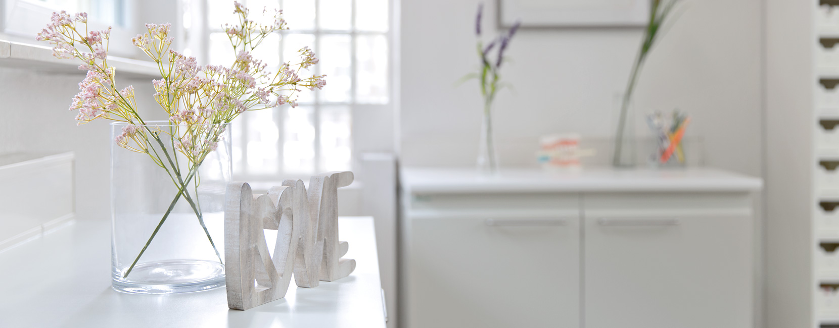 Holzaufsteller "LOVE" steht neben einer Glasvase mit zarten rosa Blüten auf einer hellen Arbeitsfläche in einem freundlichen, minimalistisch eingerichteten Raum mit Schrank und Fensterlicht.