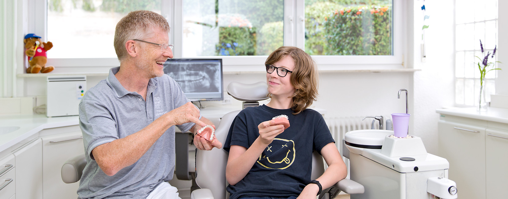 Dr. Süßenberger zeigt einem Teenager ein Zahnmodell, der Teenager hält ein weiteres Modell und lächelt; helle, moderne Praxis mit Behandlungsstuhl, Röntgenbildschirm und Fensterblick.