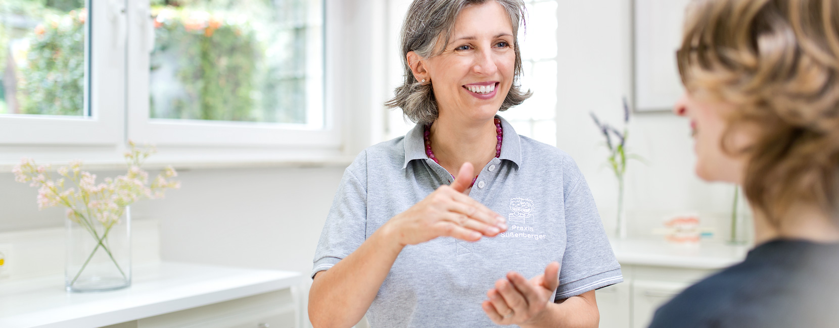 Dr. Süßenberger lächelt und gestikuliert, erklärt etwas einer Patientin; helle, freundliche Praxis mit Fenster und Blumen, Schriftzug auf dem Poloshirt: "Praxis Süßenberger".
