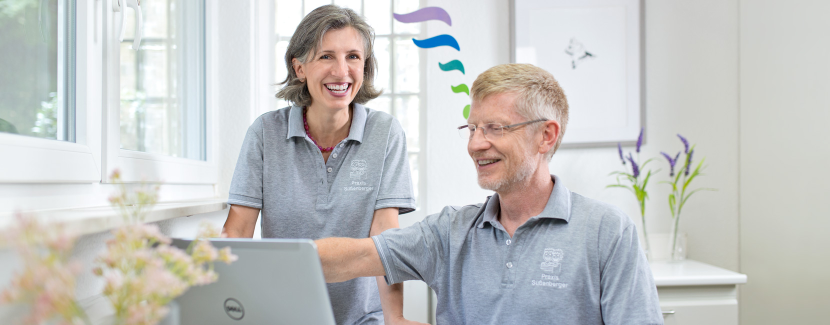 Unsere Zwei Kieferorthopöden arbeiten gemeinsam am Laptop; einer zeigt auf einen Bildschirm. Helle Praxis mit Fenster und Blumen. Poloshirts mit Aufschrift: "Praxis Süßenberger". Laptop: "Dell".